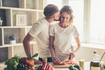 Beautiful young couple is talking and smiling while cooking healthy food in kitchen at home. Man is kissing his girlfriend in cheek